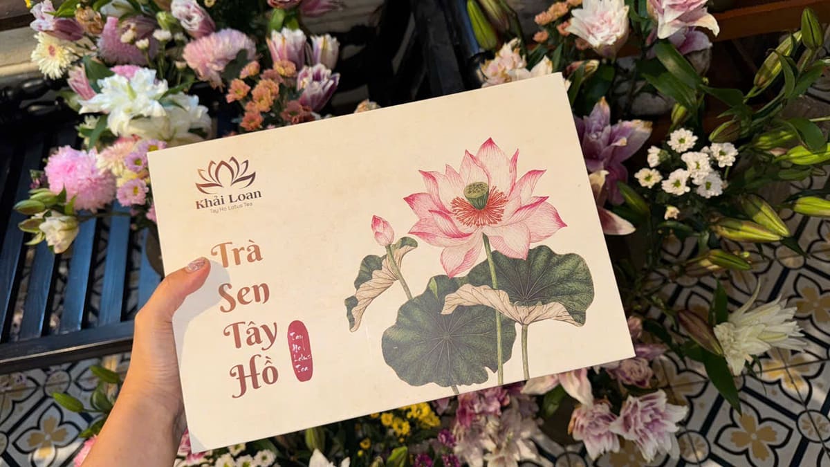 Bàn tay cầm hộp trà sen Tây Hồ in hình hoa sen giữa các bó hoa tươi